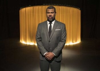 Jordan Peele stands in a darkened studio wearing a grey suit, photographed for The Twilight Zone