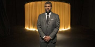 Jordan Peele stands in a darkened studio wearing a grey suit, photographed for The Twilight Zone