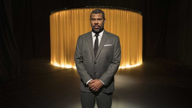 Jordan Peele stands in a darkened studio wearing a grey suit, photographed for The Twilight Zone
