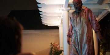 Big Baby slasher villain stands on a porch at night in a bloodstained onesie, wielding an axe, in Spider One's 2026 horror film.