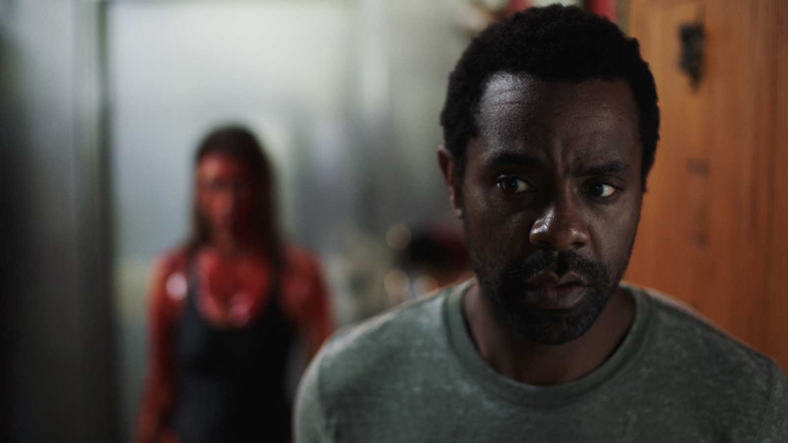 Adam Lewis looks over his shoulder at a blurred, blood-covered figure in a doorway in Spider One's meta slasher Big Baby.