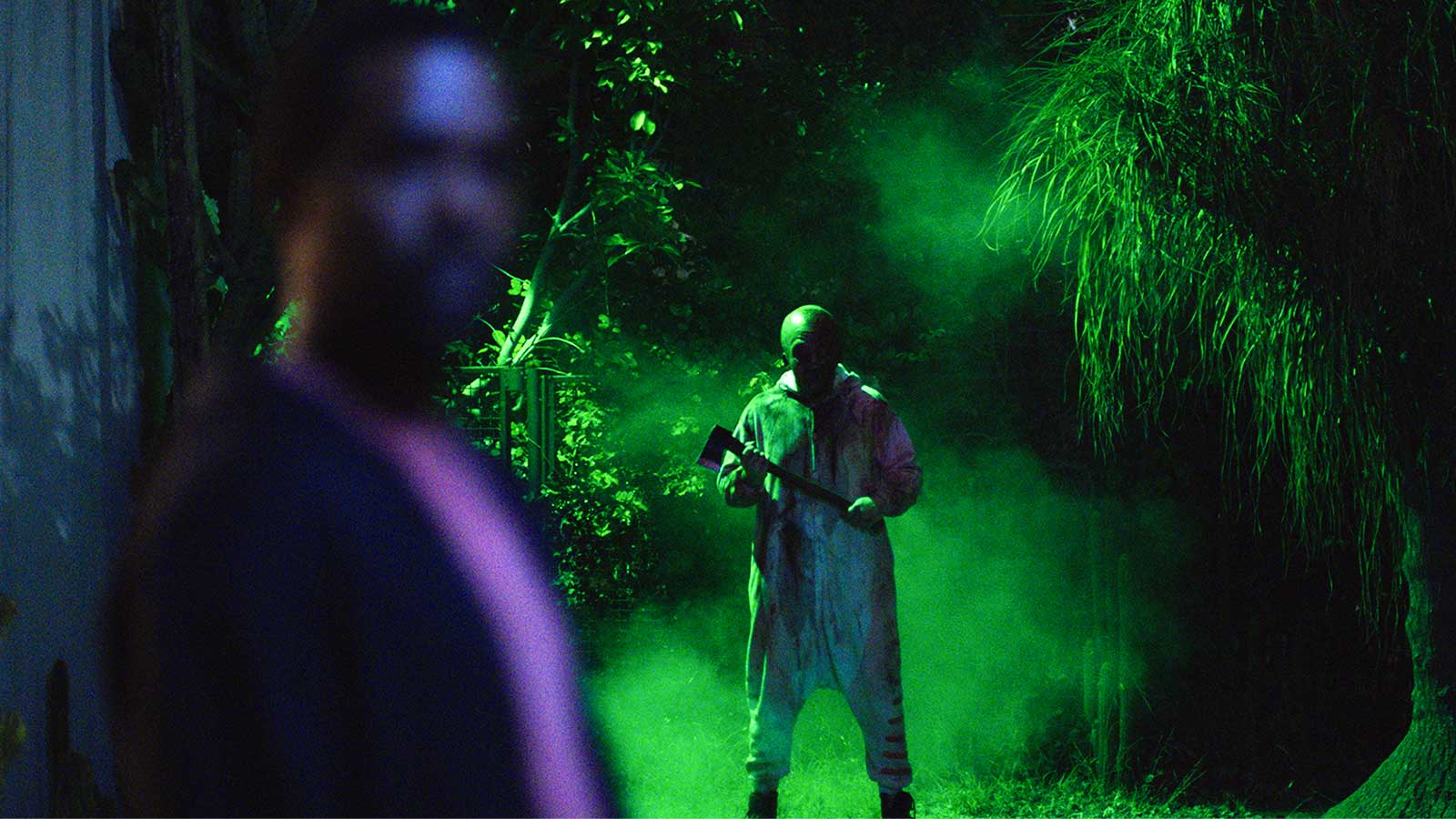 Big Baby's masked killer looms in a fog-filled, green-lit garden behind an unaware man in Spider One's 2026 horror film.