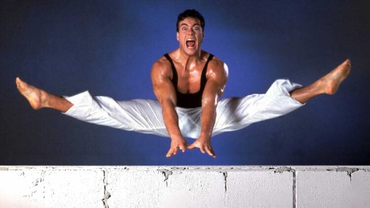 Jean-Claude Van Damme mid-air performing a split jump over a wall in a promotional photo